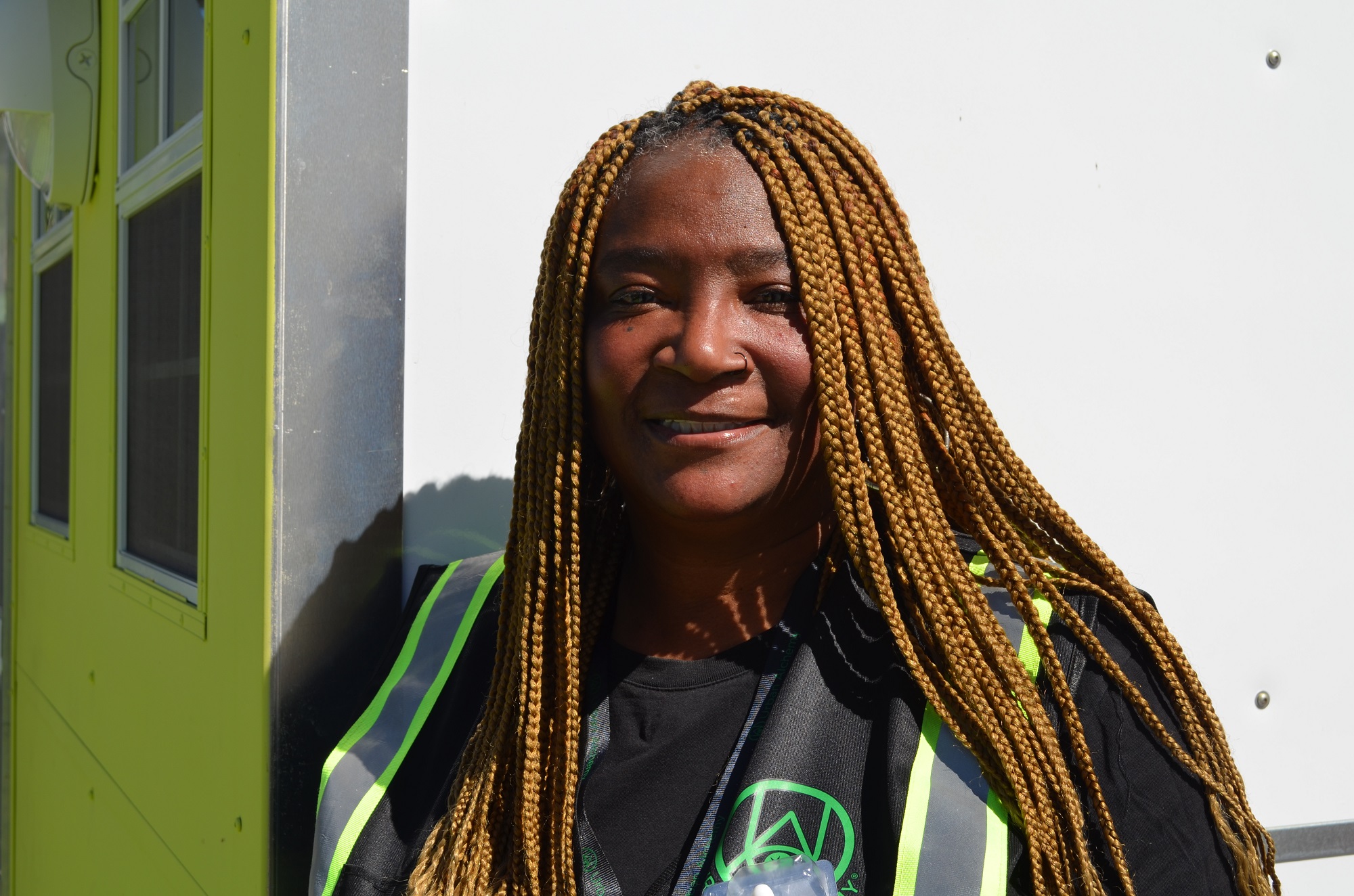 A smiling woman with long braided hair, wearing a brightly colored construction vest over a dark shirt.