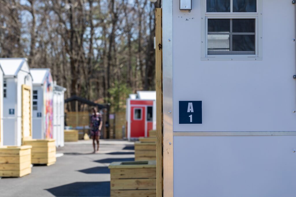 Two rows of Pallet shelters in Boston, Massachusetts. One shelter in the foreground is in focus. A person walking in the background in between the rows is slightly blurry.