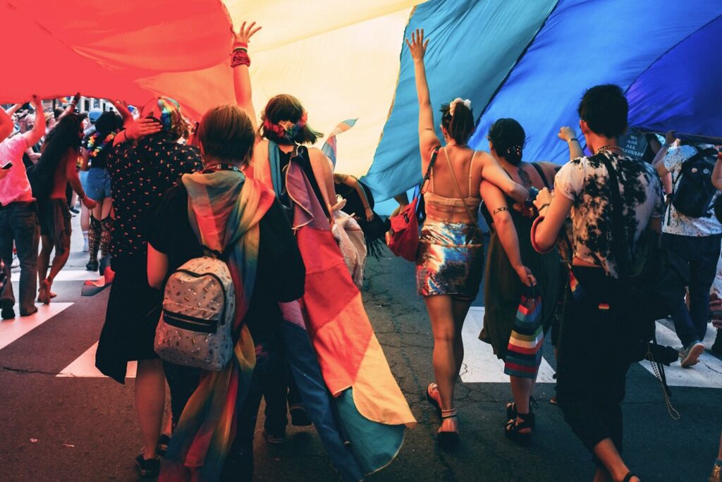 A group of people seen from behind walking underneath a large rainbow flag.