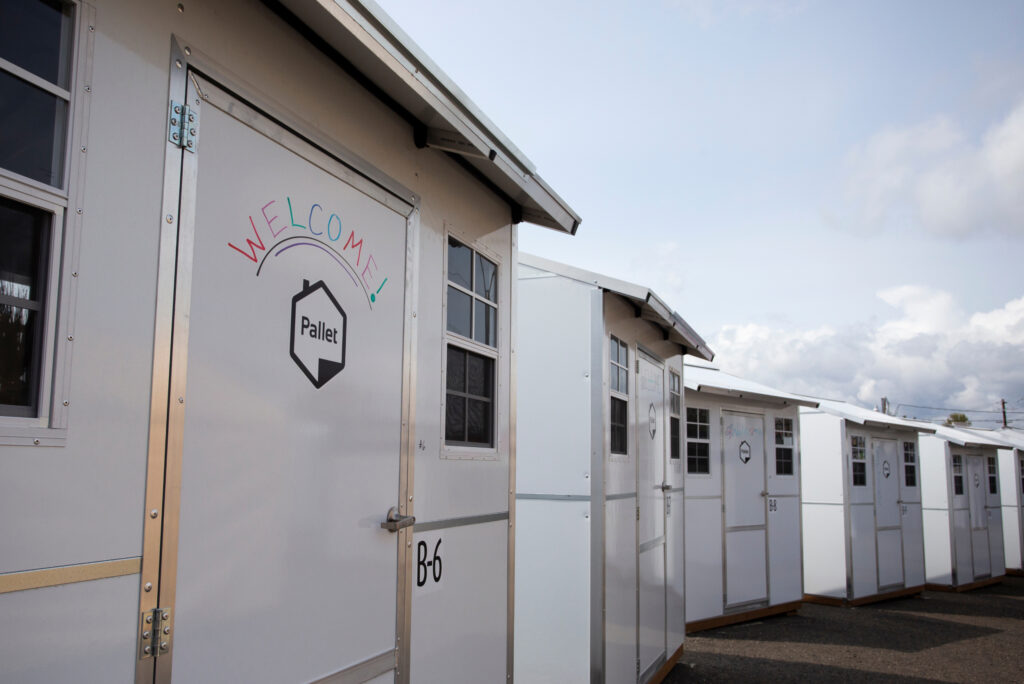 A row of five Pallet shelters. The phrase welcome is written on one of the doors.