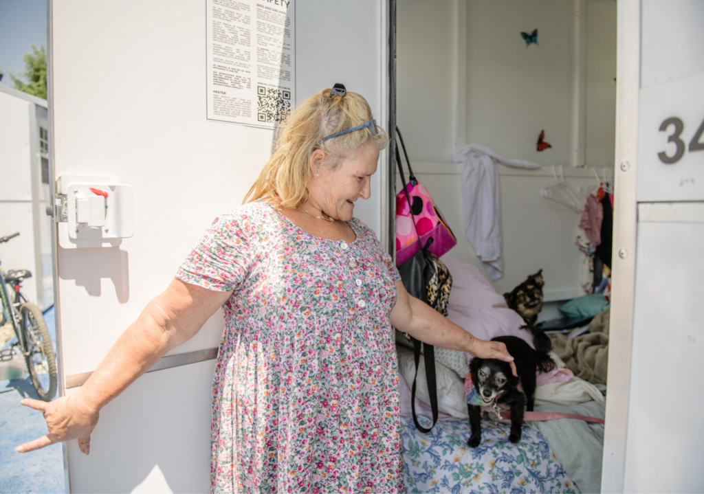 Lynette holds the door open to her Pallet shelter while petting one of her two dogs.