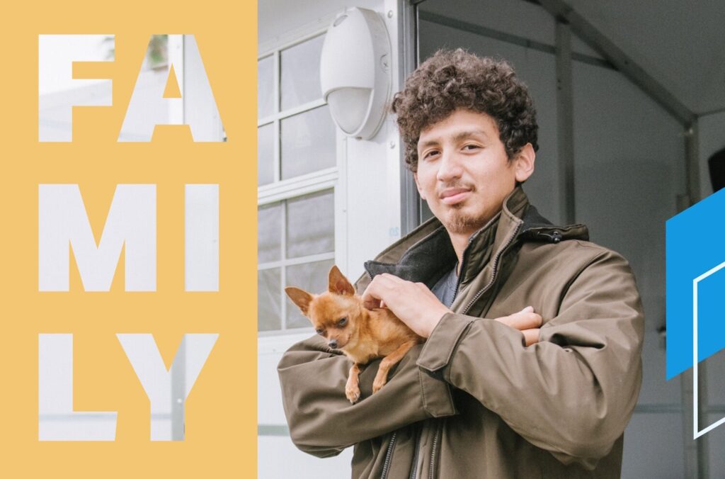 A man in a heavy-duty jacket smiles as he pets a small dog in his arms.