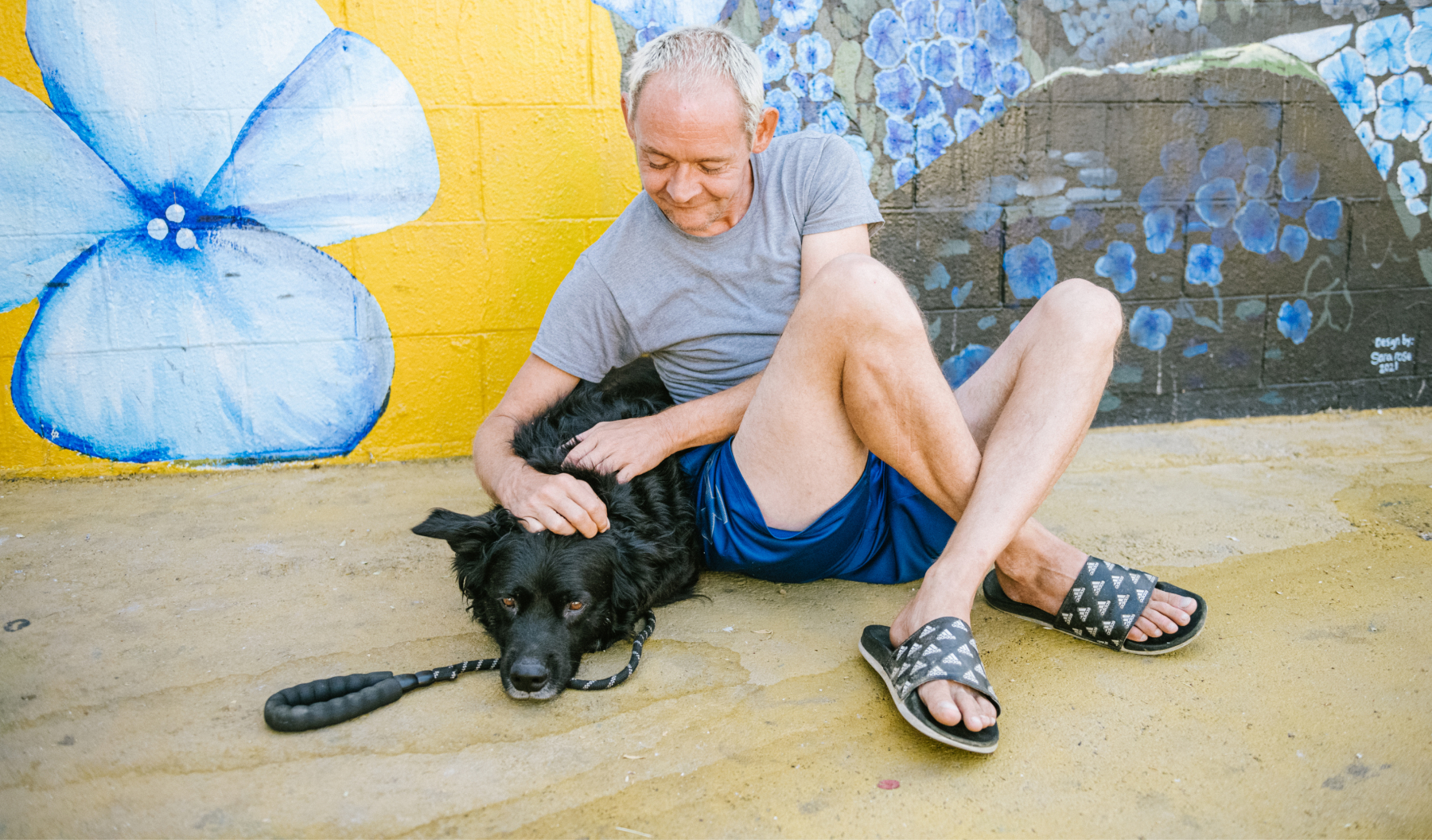 A man sits against a colorful mural while petting his dog.
